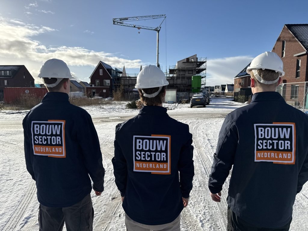Drie vakmensen van Bouwsector Nederland met witte helm kijken over een besneeuwde straat naar nieuwbouw met een bouwkraan